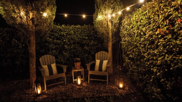 Un espacio al aire libre con dos sillas Adirondack, una mesita y faroles, rodeado de árboles y luces colgantes.