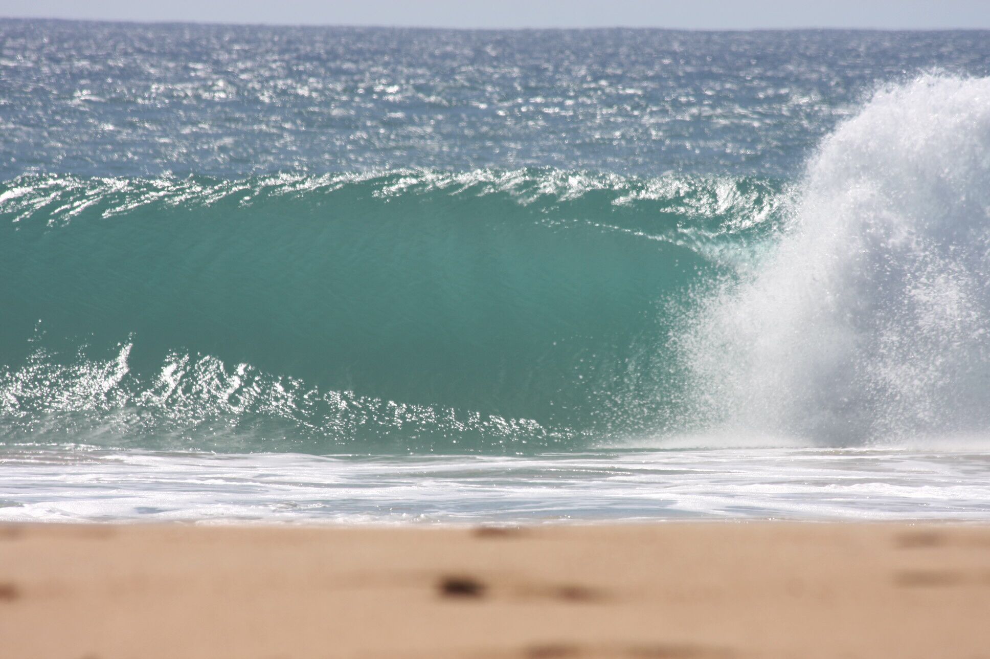 A large wave in the ocean with a sandy beach in the foreground.