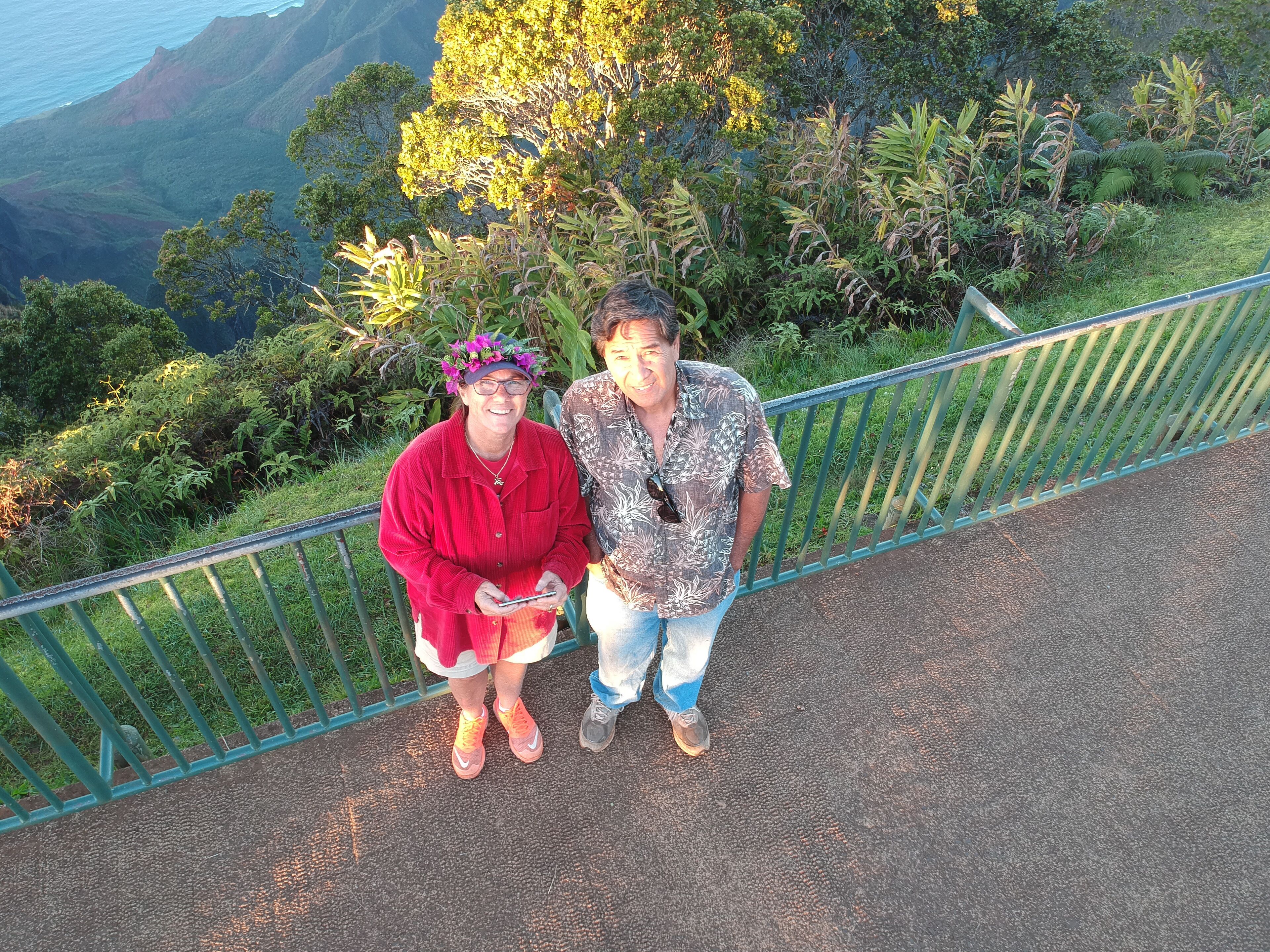 Two people standing on a pathway with a railing, surrounded by lush greenery and a mountainous backdrop.