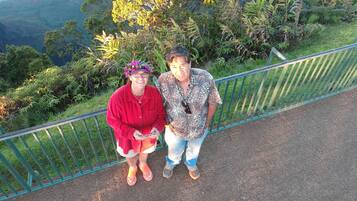 Two people standing on a pathway with a railing, surrounded by lush greenery and a mountainous backdrop.