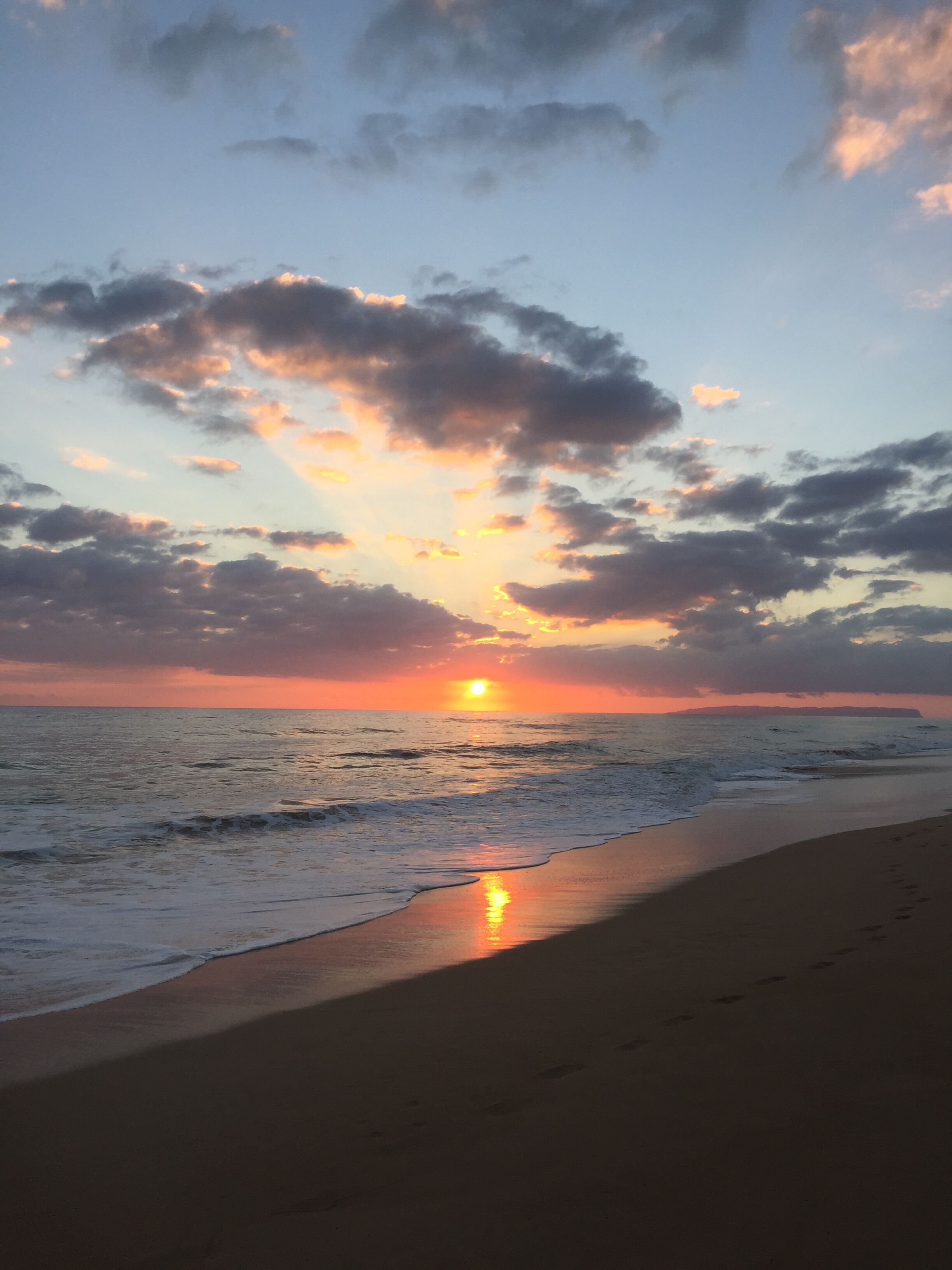 A beach at sunset with the sun setting over the horizon, clouds in the sky, and waves crashing onto the shore.