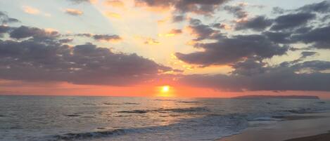 A beach at sunset with the sun setting over the horizon, clouds in the sky, and waves crashing onto the shore.