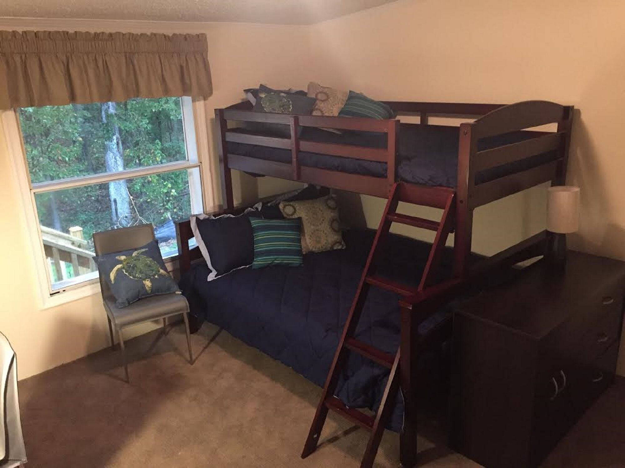 A bunk bed with a desk and chair, a window with a view of trees, and a small desk with drawers.