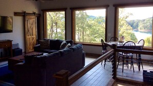 Un salón con sofá, televisor, mesa de madera con sillas y ventanas amplias con vistas a árboles y un lago.