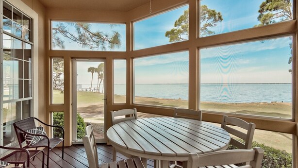 A covered outdoor seating area with a table and chairs, large windows overlooking a beach and ocean.