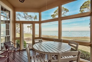 Un espacio de estar techado al aire libre con una mesa y sillas, amplias ventanas con vista a una playa y el océano.