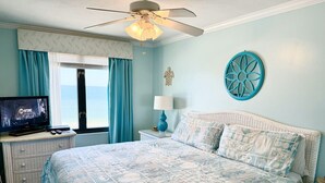 Ein Schlafzimmer mit Bett, Fernseher, Deckenventilator, einem Fenster mit Vorhängen und einem Wanddekor.