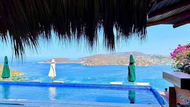 Ein Pool mit Blick auf das Meer und die Hügel, Sonnenschirme und ein Reetdach.