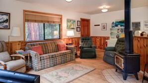 A cozy living room with a plaid sofa, a green armchair, and a black wood-burning stove. - (1S) Cecil's Cabin at The Redwoods In Yosemite (Wawona)
