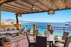 Una terrassa amb mobles de vusta, una taula preparada per dinar i vista al mar i un vaixell de creuer.
