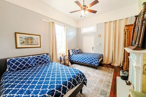 Ein Schlafzimmer mit zwei Betten, einem Deckenventilator, einem Fenster mit Vorhängen und einem gerahmten Bild an der Wand.