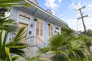 A two-story building with a blue facade, white shutters, and a small porch with potted plants.