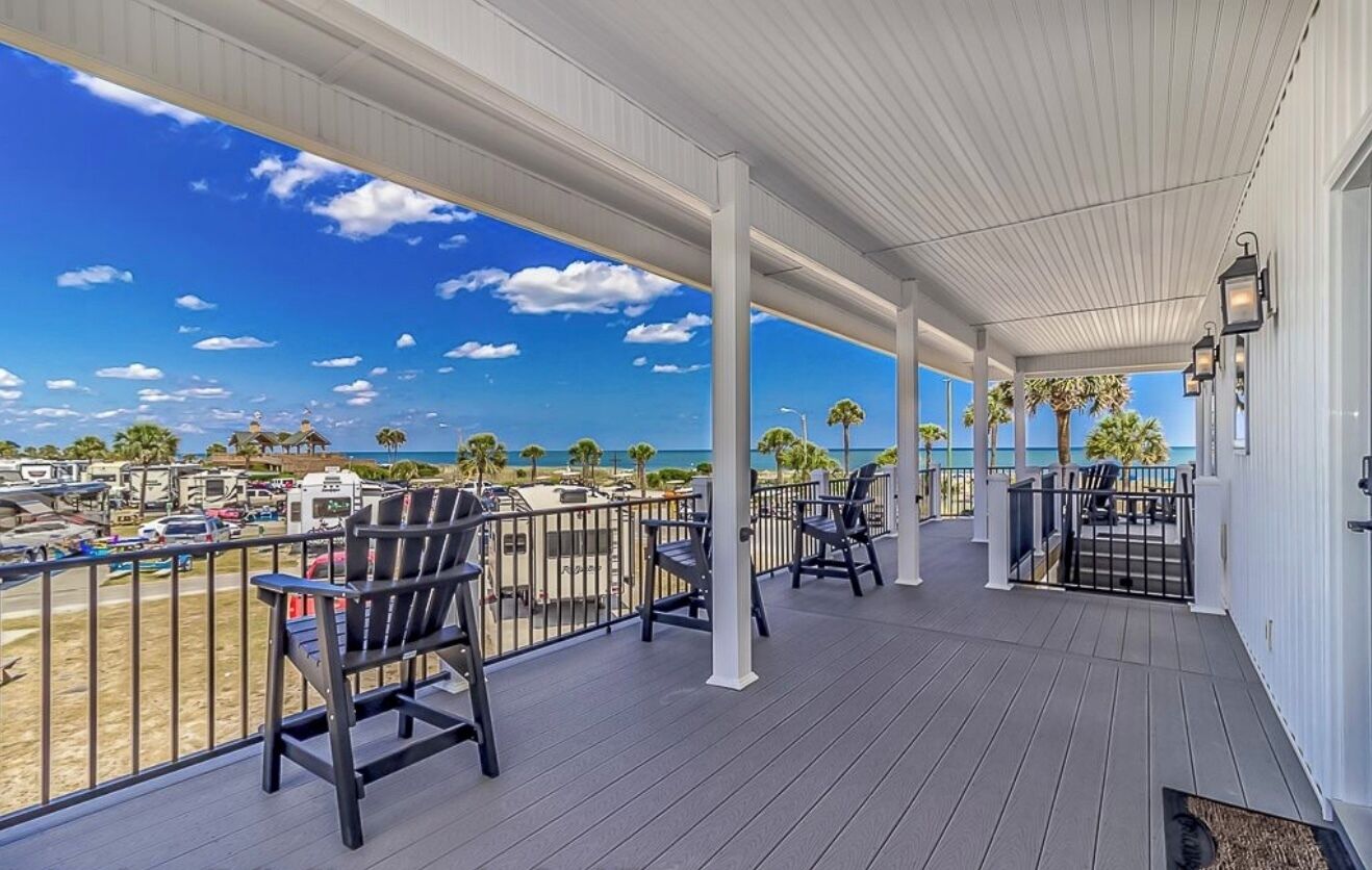 A covered outdoor area with chairs and tables, overlooking a parking lot and beach with palm trees.
