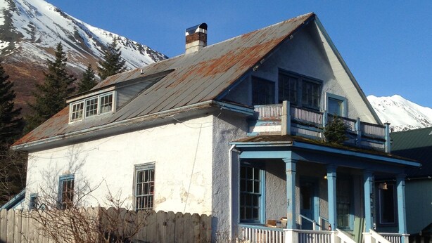 A two-story house with a balcony and a porch, surrounded by a wooden fence and a mountain backdrop. - The 1905 Government. cable Office: Historic Charm, views, location Downtown (Seward)