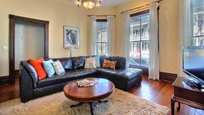A living room with a sectional sofa, a coffee table, a chandelier, and a large window with curtains. - Big Blue House - Lower Unit - Near Forsyth Park - Sleeps 8 (Savannah)