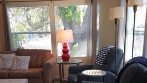 Un salon avec un tapis à rayures, un canapé marron, un fauteuil bleu, une petite table ronde avec une lampe rouge et une grande fenêtre avec des stores.
