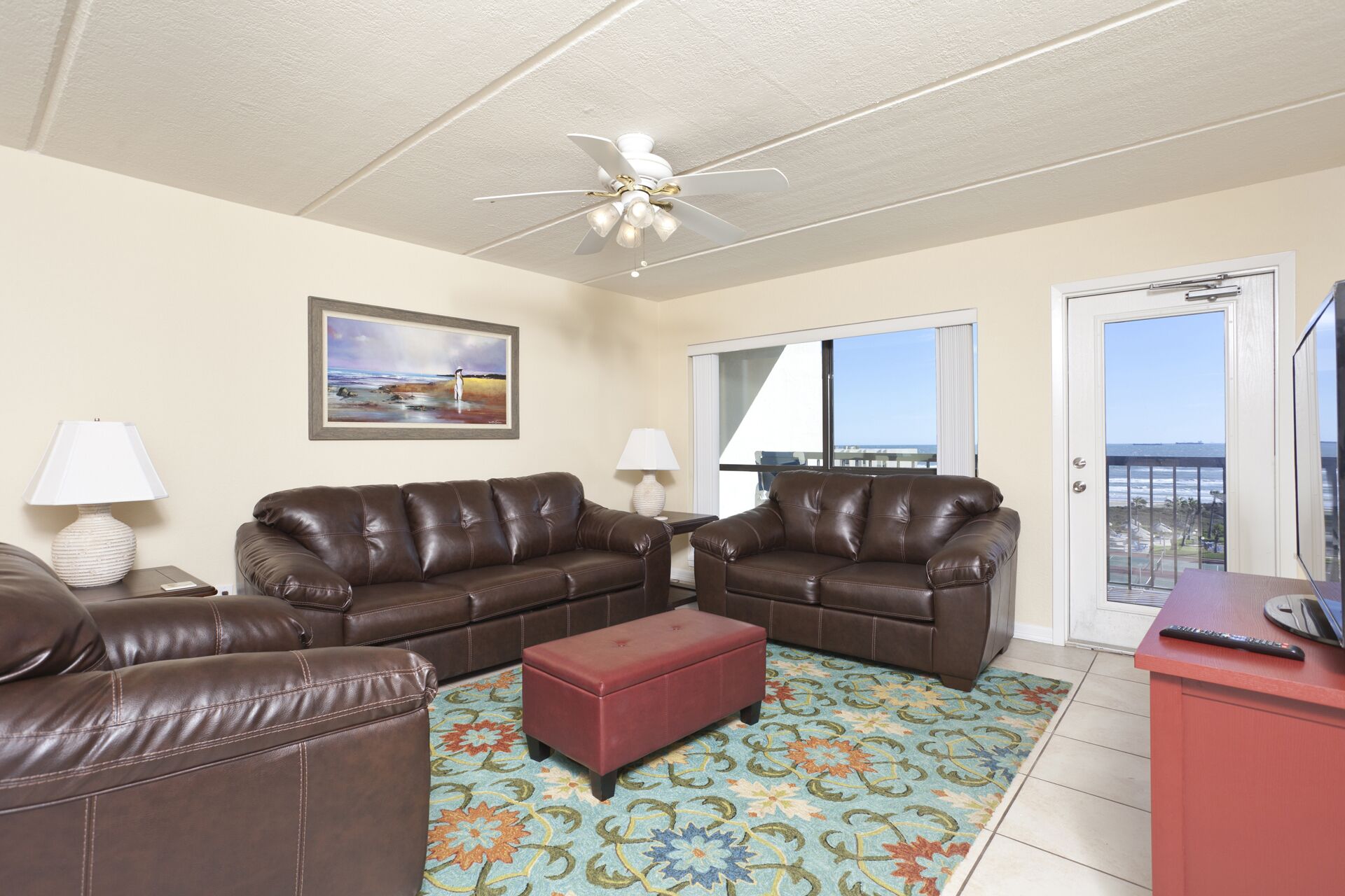A living room with a brown leather sofa set, a red ottoman, and a patterned rug. There is a ceiling fan, a painting on the wall, and a view of the ocean through a sliding glass door.