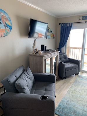 Ein Wohnzimmer mit grauem Sofa, einem kleinen Tisch mit Fernseher und einem Balkon mit blauen Vorhängen.