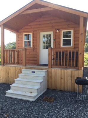 A wooden cabin with a front porch, a set of stairs leading to the door, and a grill outside.