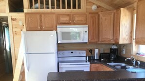 A kitchen with wooden cabinets, a microwave, a stove, and a sink. There are two black stools and a trash can. -   Little fishing cabin by the lake “Come catch some memories “ (Scottsboro)