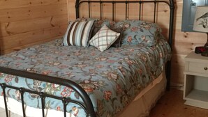 A wooden cabin room with a metal bed frame, floral bedding, and a bedside table with a lamp. -   Little fishing cabin by the lake “Come catch some memories “ (Scottsboro)