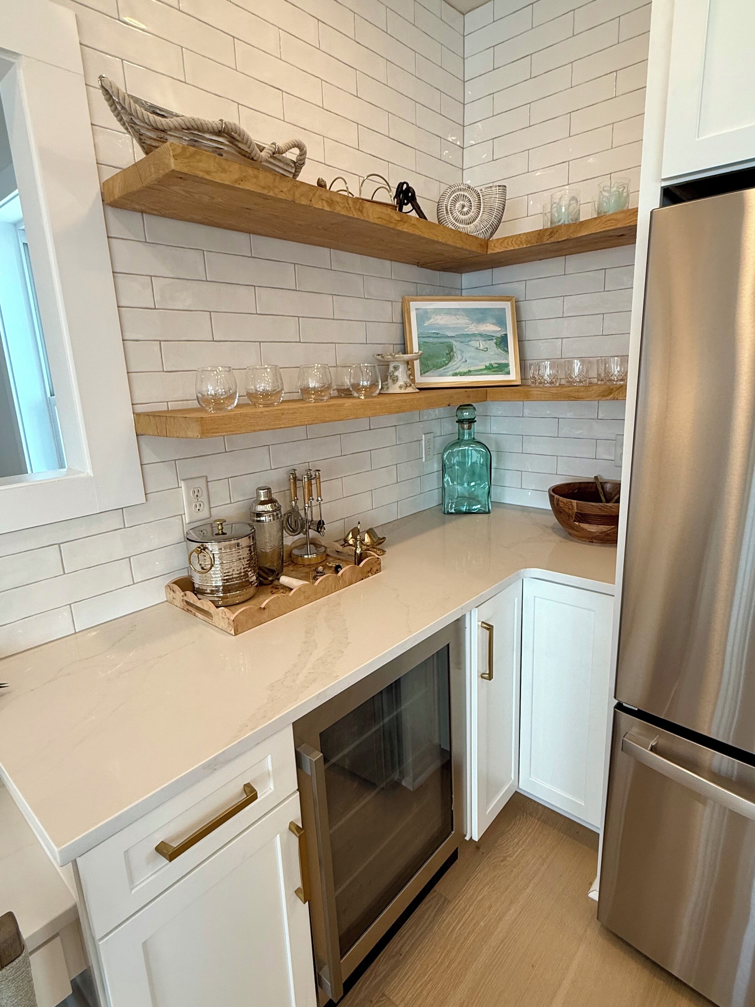 A modern kitchen with white cabinetry, a stainless steel refrigerator, and wooden shelves holding glassware and a painting.
