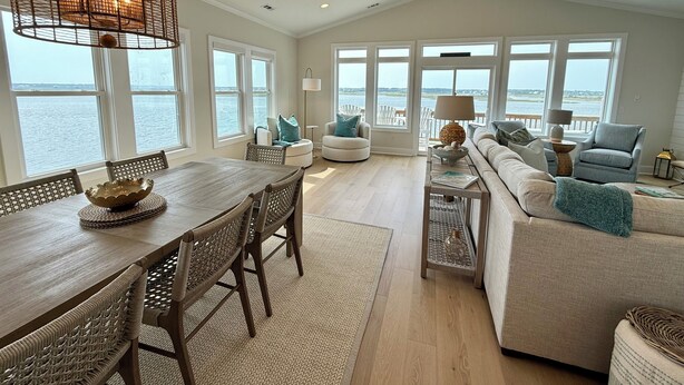A spacious living room with a large window overlooking the water, a dining area with a wooden table and wicker chairs, and a cozy seating area with a sofa and armchairs. - Best Views on the Island! Oceanfront, Sunsets, Boat dock, Elevator, pool, fish! (Topsail Beach)