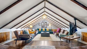 Een loft-achtige woonkamer met een hoge plafond, houten balken, een zithoek met banken en een salontafel, en een groot vloerkleed.