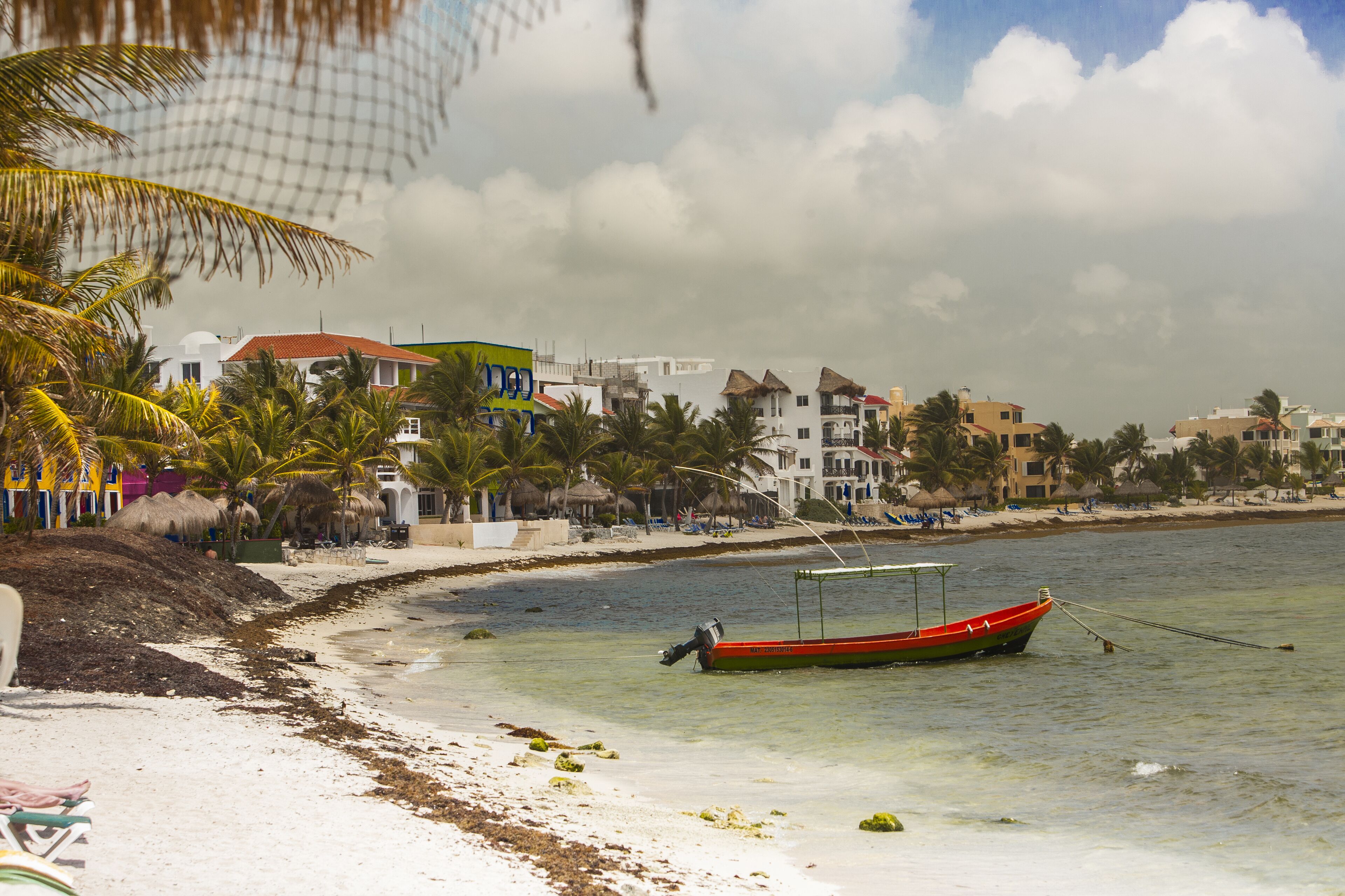 Ein Strand mit einem roten Boot, das in der Nähe des Ufers vor Anker liegt, umgeben von Palmen und Gebäuden.