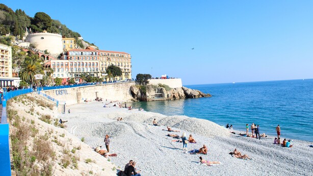 Une ville côtière dotée d’une plage de galets, d’un bâtiment qui ressemble à un château et d’un ciel bleu azur.