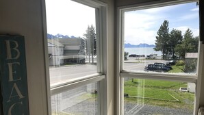 Uitzicht vanuit een raam, met een 'BEACH' bord aan de linkerkant, een parkeerplaats met auto's en een weg die naar een gebouw leidt, met bergen op de achtergrond.