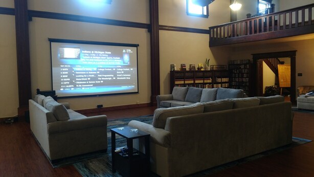 A spacious living room with a large screen displaying sports scores, a comfortable sofa set, a coffee table, and a bookshelf. - Knights of Pythias Loft Downtown Albany, Oregon 2400 Sq Ft Of Space (ALBANY)