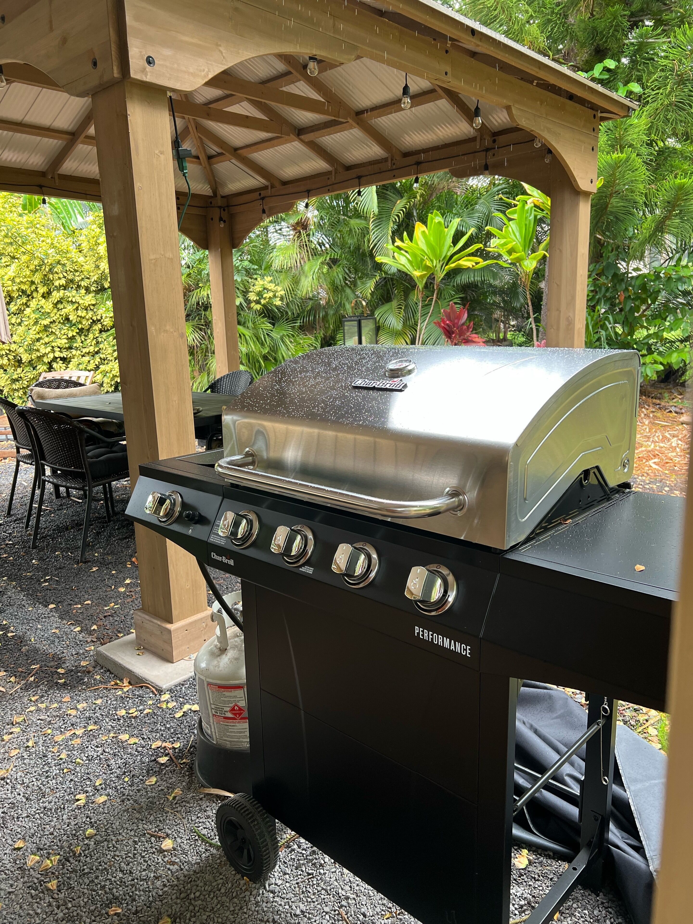 Char-Broil-merkkinen, nelipolttimoinen grilli, jossa on kaasupullo ja metalliritilä. Grilli on sijoitettu puisen pergola-rakenteen alle puutarhaan, jossa kasvaa monenlaisia kasveja.