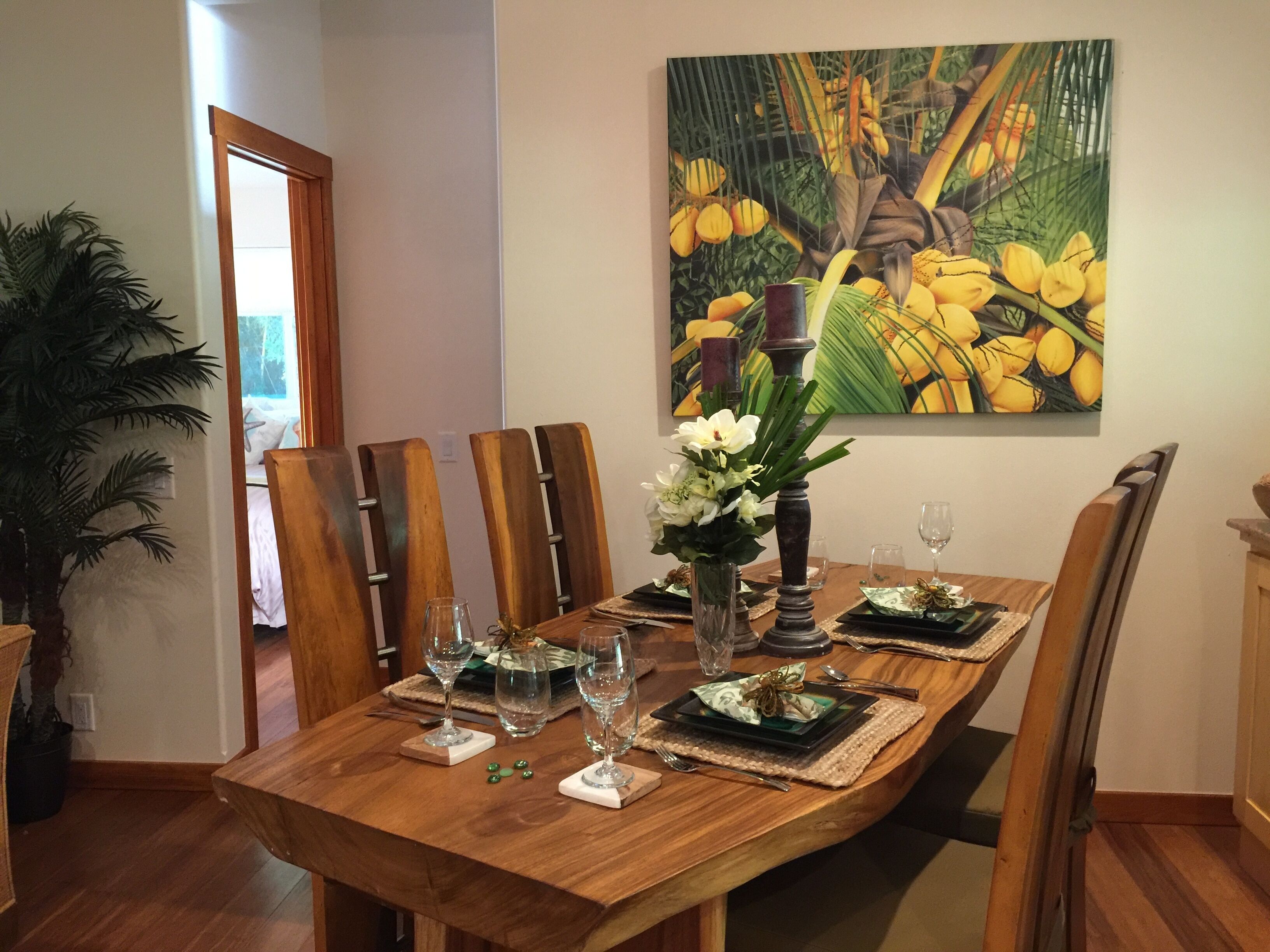 Uma sala de jantar com uma mesa de madeira posta para quatro pessoas, rodeada por cadeiras. Um quadro grande com frutas amarelas está pendurado na parede.