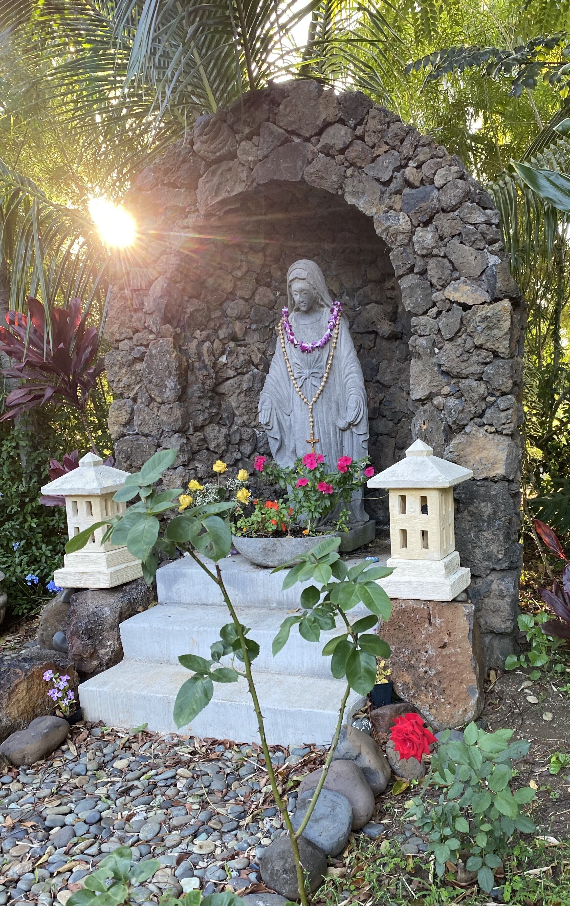 A statue of a religious figure under a stone archway, surrounded by flowers and plants.