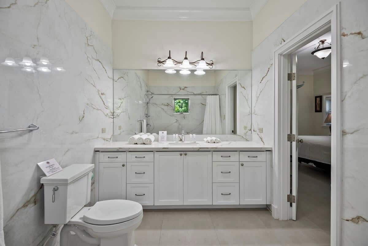 Un baño con pared de mármol, inodoro blanco, lavamanos con espejo y zona de ducha.