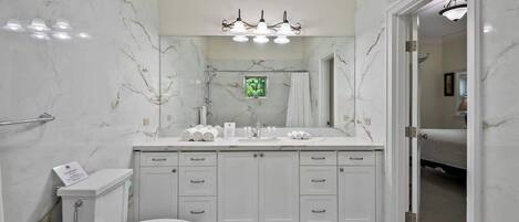 Un baño con pared de mármol, inodoro blanco, lavamanos con espejo y zona de ducha.