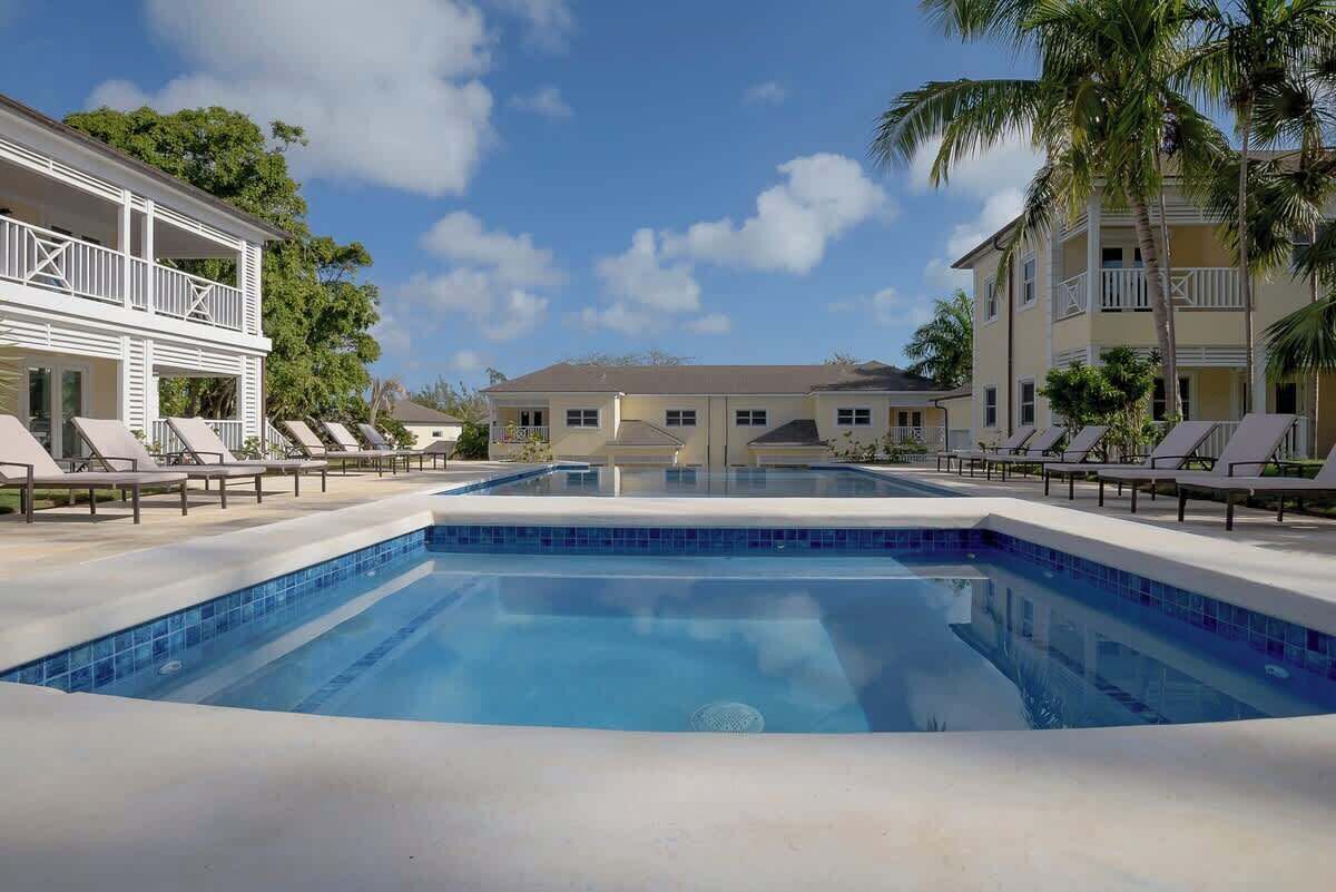 Una piscina con sillas de descanso, un edificio de dos pisos con balcones y palmeras.
