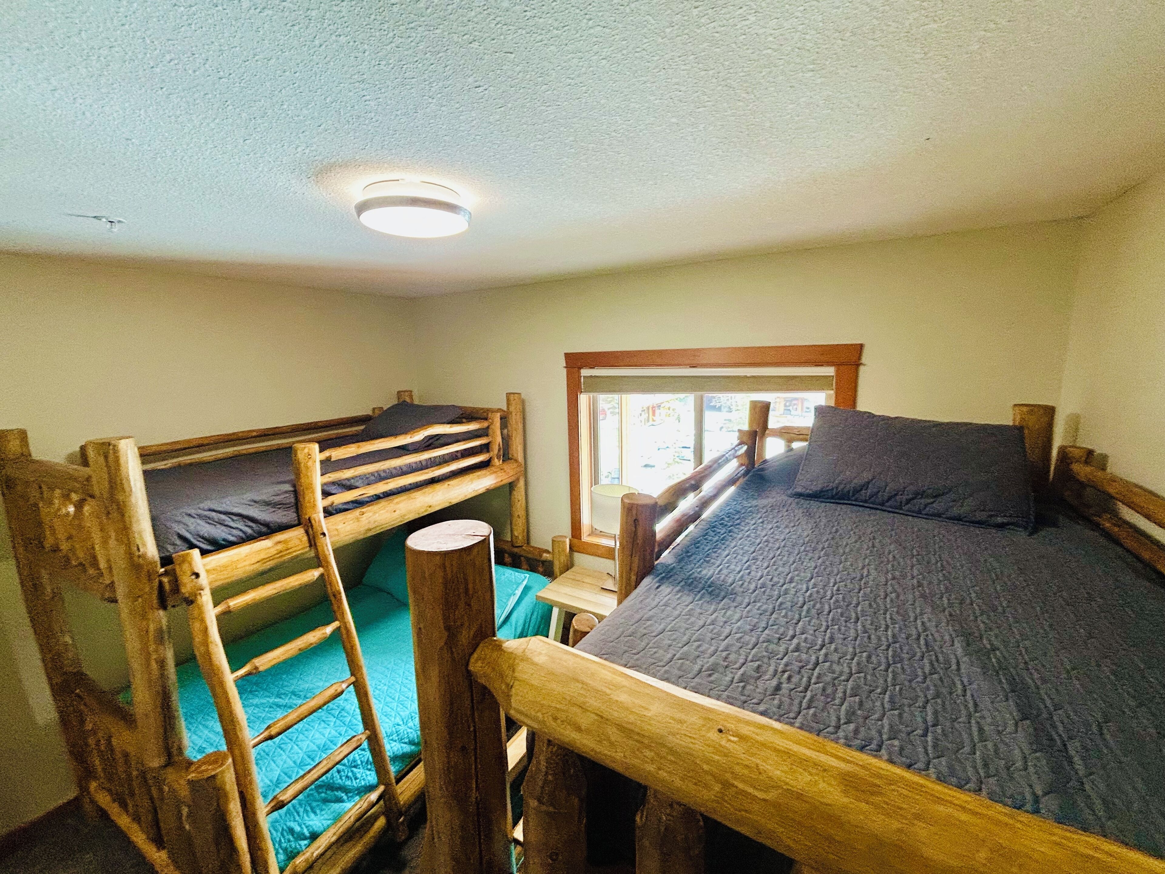 A dormitory room with two bunk beds, a wooden ladder, a window with a view, and a ceiling light.