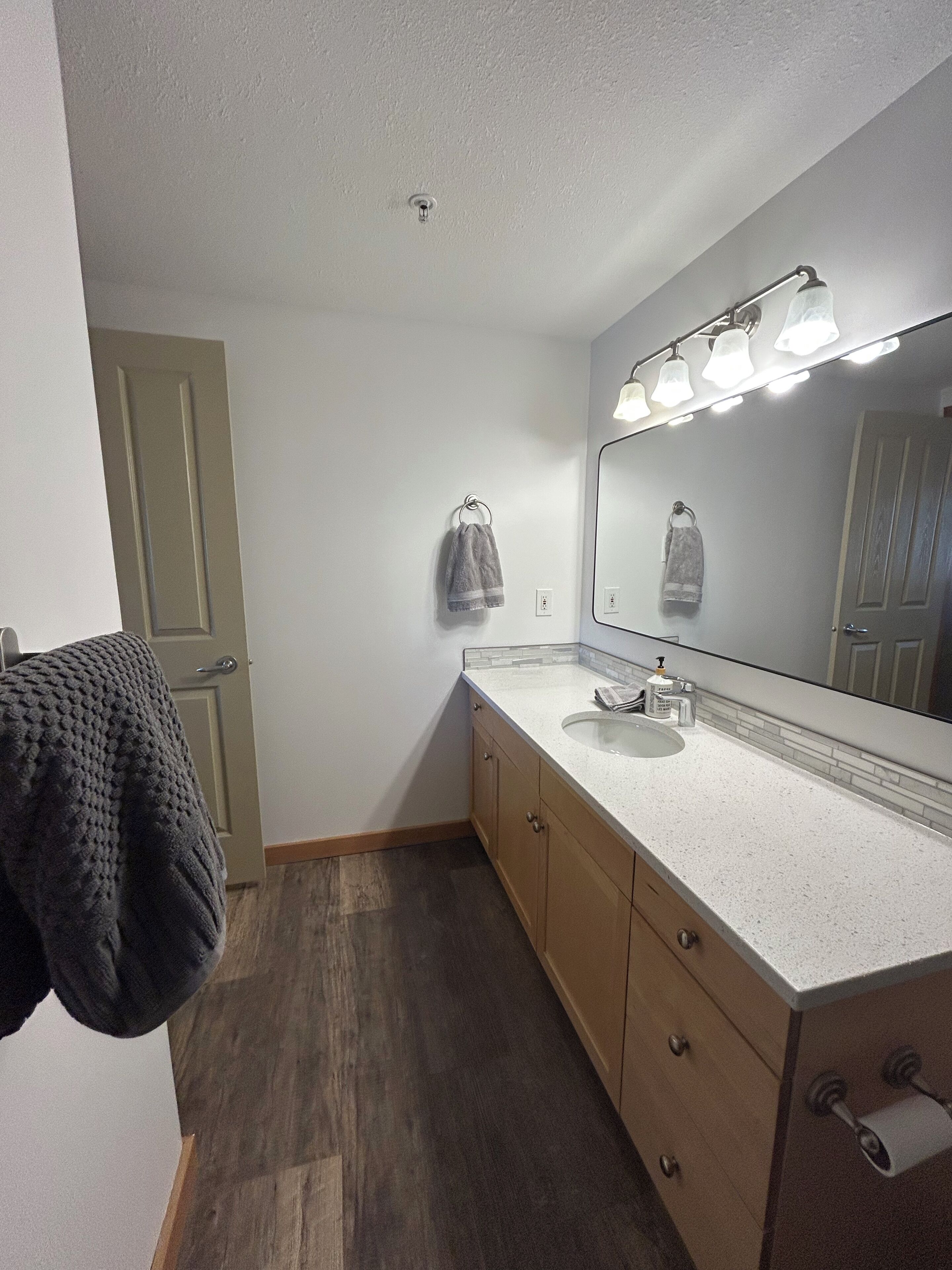A bathroom with a white sink, a mirror with lights, wooden cabinets, and a towel hanging on the wall.