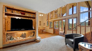 A spacious living room with a large flat-screen TV, wooden shelves with decorative items, a fireplace, and a view of the outdoors through large windows. - Large private lot.  Beautiful cabin with a stream and trout pond!  (Lead)