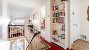 Een ruime hal met een trap die naar een balkon leidt, een grote boekenkast vol boeken, en een deur met het nummer 68.