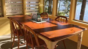 Un espace repas avec une table et des chaises en bois, un luminaire suspendu et de grandes fenêtres donnant sur un plan d’eau.