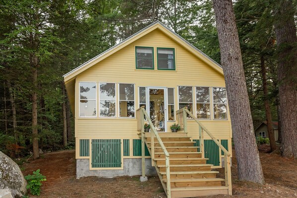 A small, single-story wooden cabin with a steeply pitched roof, large windows, and a wooden staircase leading to a front porch.