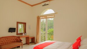 A bedroom with a bed, a sofa, a desk, a mirror, and a window with a view of greenery. - OYO Life 414 Loji Rejo Guest House (Malang)