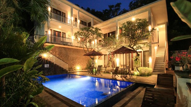 A swimming pool with a stone border, surrounded by lounge chairs and umbrellas, and a staircase leading to a balcony. - Dewi Villa Padangbai Hotel (Padangbai)