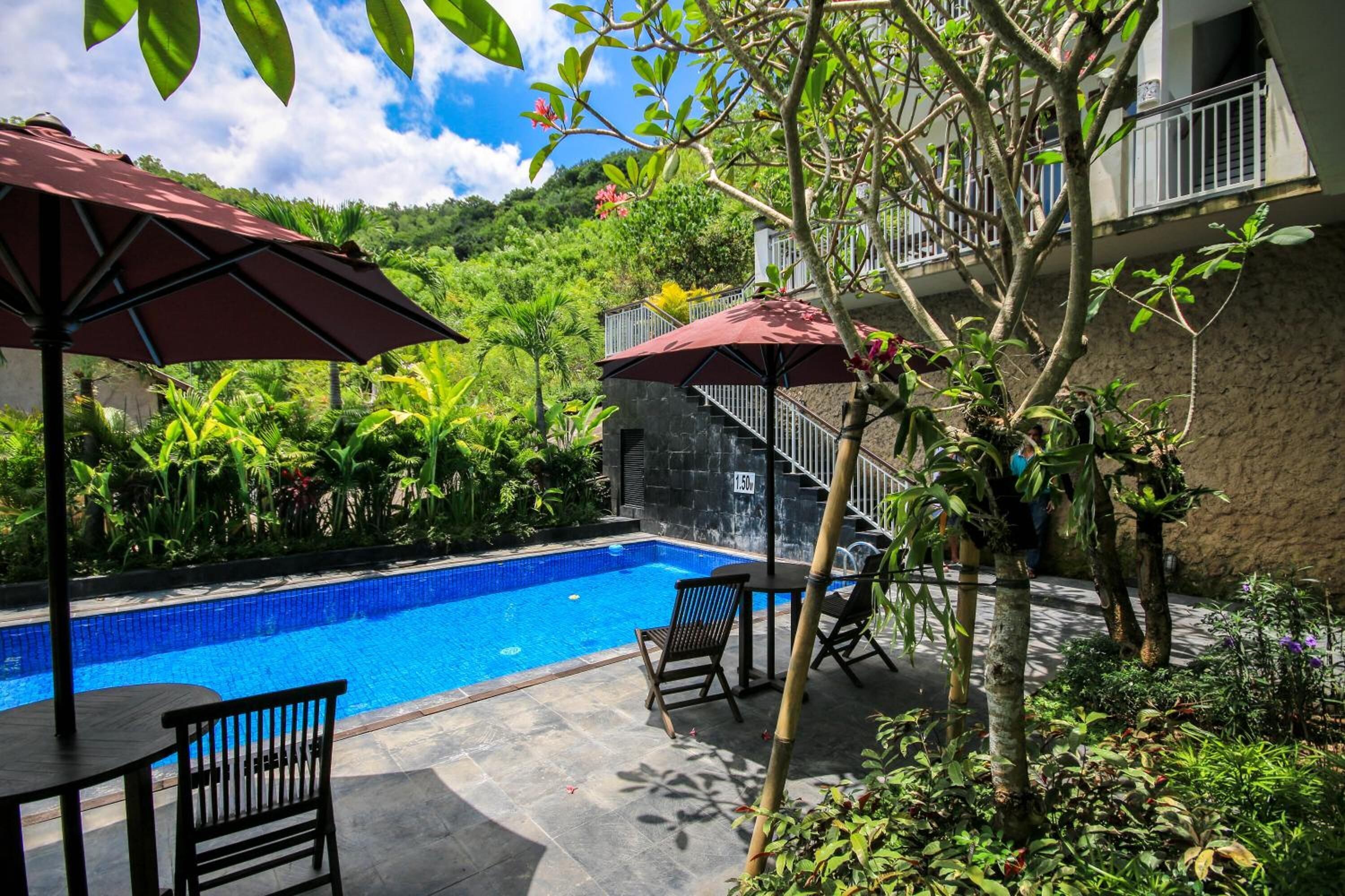A swimming pool with a stone wall, surrounded by lush greenery and two outdoor seating areas with umbrellas.
