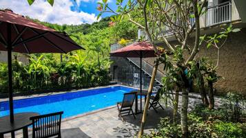 A swimming pool with a stone wall, surrounded by lush greenery and two outdoor seating areas with umbrellas.