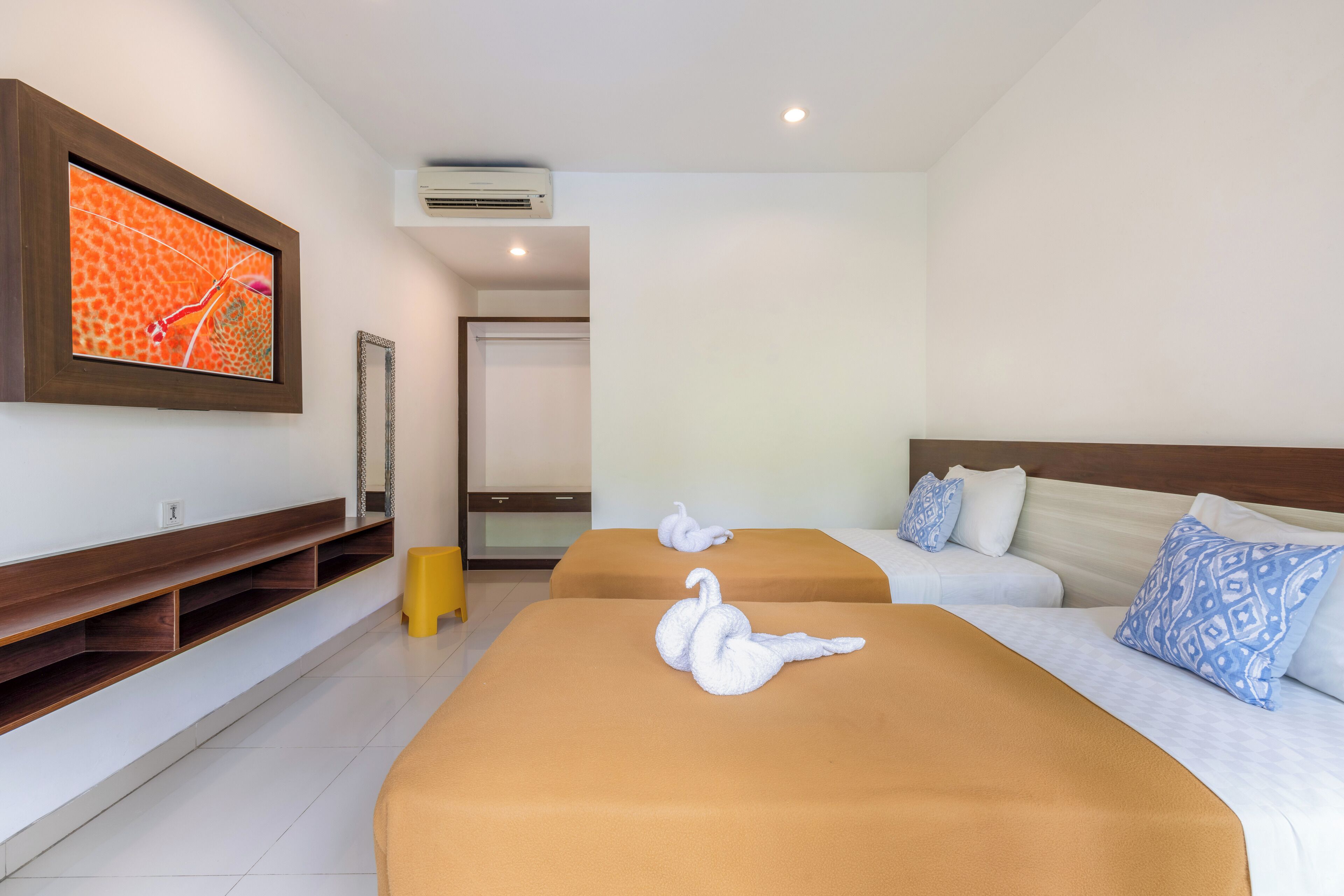 Una habitación de hotel con dos camas, un televisor montado en la pared y un taburete amarillo pequeño.