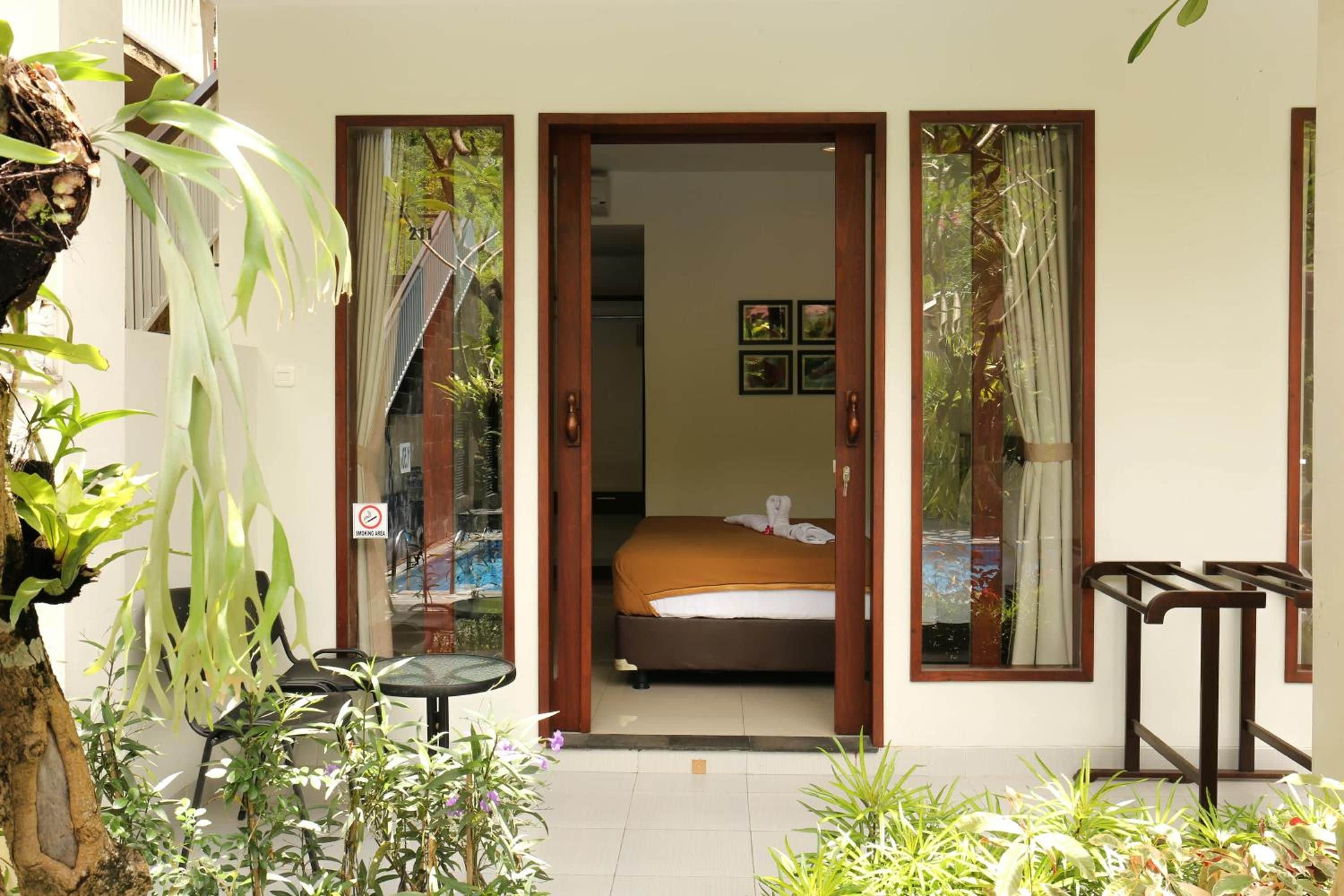 A hotel room with a bed, a small table, and a chair. There are plants outside the room.
